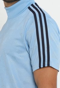 Ljusblå kortärmad sporttröja med subtila geometriska mönster och tre svarta ränder som löper ner från axeln till ärmen.