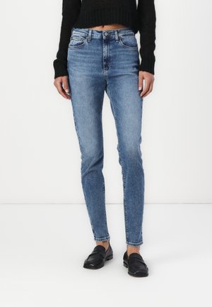 Frau trägt blaue Skinny-Jeans mit mittlerer Bundhöhe, schwarzen kurzen Pullover und schwarze Loafers, steht vor weißem Hintergrund.