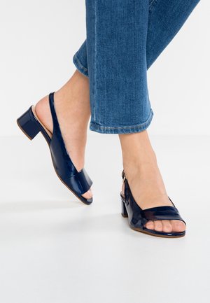 Pies con sandalias de tacón ancho en bloque, de charol azul oscuro y diseño slingback, combinadas con jeans vaqueros azules recortados sobre suelo blanco.