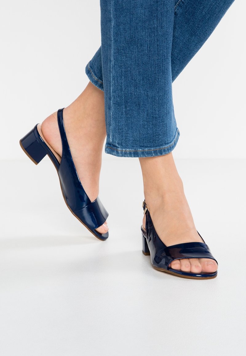 Pieds portant des sandales à talons blocs à brides arrière en cuir verni bleu foncé associées à un jean denim bleu raccourci sur un sol blanc.