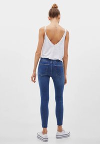 Kvinna med hår i en knut iklädd en vit rygglös linne, blå tajta jeans och vita platåskor som står mot en neutral bakgrund.