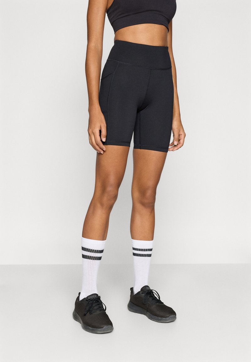 Μαύρο αθλητικό σορτς με ψηλή μέση και ραφές επίπεδης ραφής, φορεμένο με λευκές κάλτσες που έχουν μαύρες ρίγες και μαύρα παπούτσια.