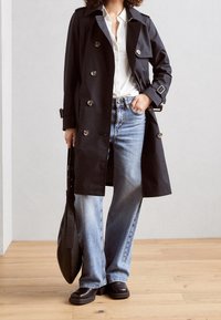 Svart trenchcoat med dubbelknapad design, stora knappar och bälte. Bärs över en vit skjorta, ljusblå vida jeans och svarta ankla stövlar.