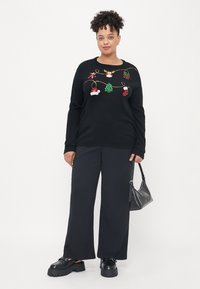 Pulover negru cu broderie colorată și festivă, prezentând decorațiuni și cadouri; asortat cu pantaloni largi negri și pantofi masivi negri, purtând o geantă.