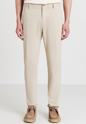 Homme portant un pantalon beige droite et des chaussures en daim marron à lacets, debout avec les mains détendues sur les côtés contre un fond blanc.