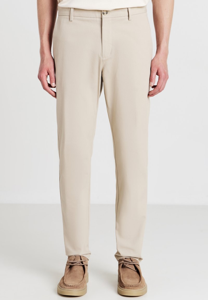 Homme portant un pantalon beige droite et des chaussures en daim marron à lacets, debout avec les mains détendues sur les côtés contre un fond blanc.
