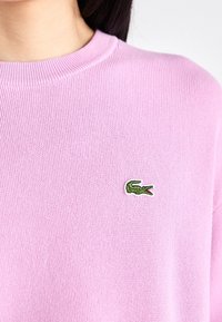 Pull en maille rose clair avec un col rond et un logo croco vert brodé sur la poitrine. Texture douce et coupe standard.