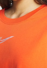 Orange bomullströja med rund halsringning och ribbad krage, med en vit logotyp i en dynamisk design på framsidan. Slät textur.