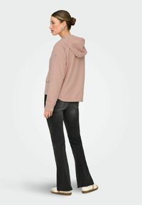 Femme aux cheveux attachés en chignon, portant une veste à capuche rose, un jean évasé noir et des baskets blanches, debout de profil sur un fond clair.