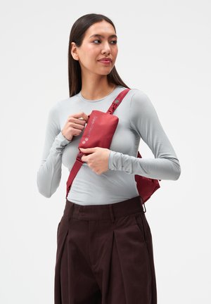 Frau mit langen dunklen Haaren, die ein graues Langarmshirt und dunkle Hosen trägt, hält eine rote Umhängetasche nahe ihrer Brust.