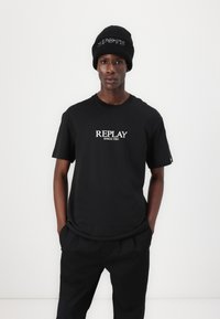 Juodas medvilnės marškinėlis su balta „REPLAY SINCE 1981“ spauda. Modelis taip pat dėvi juodą kepuraitę su kontrastuojančiu tekstu. Paprasta, laisva silueto dizaino.