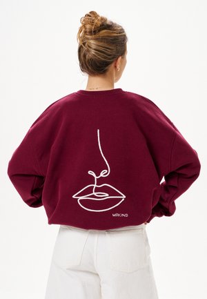 Vrouw met haar in een knot, draagend een bordeauxrode sweatshirt met een witte minimalistische lijntekening van lippen en neus op de achterkant.