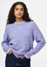 Jeune femme avec de longs cheveux bouclés, portant un pull en laine violet clair et un jean bleu foncé, debout avec les mains dans les poches.