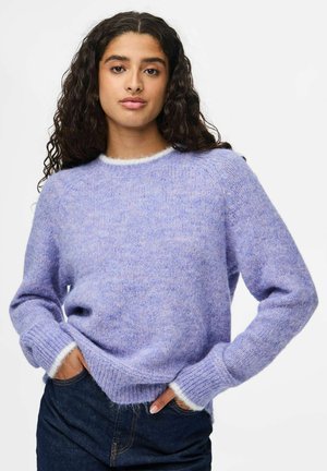 Jeune femme avec de longs cheveux bouclés, portant un pull en laine violet clair et un jean bleu foncé, debout avec les mains dans les poches.