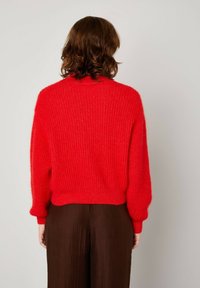 Raudonas megztinis su tekstūru, apvaliu iškirpimu ir nuleistomis pečių siūlėmis. Dėvimas su rudomis, plačiomis kelnėmis.