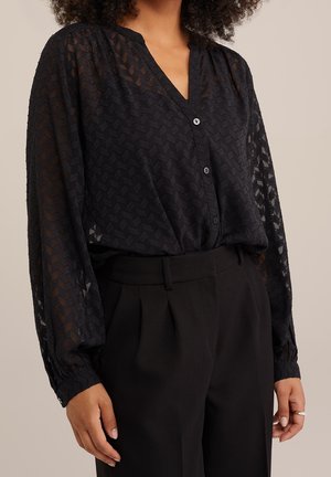 Femme portant un chemisier noir texturé à manches longues avec des boutons et un pantalon noir taille haute, debout devant un fond neutre.