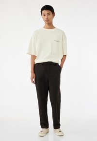 Helles gelbes oversized T-Shirt mit gesticktem Logo, kombiniert mit dunkelbraunen, taillierten Hosen und cremefarbenen Sneakers. Glatte Stoffqualität.