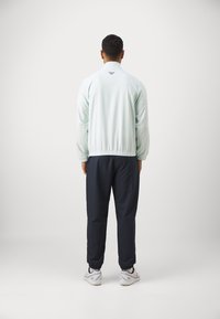 Casaco verde menta claro com tecido texturizado, punhos elásticos e um logótipo escuro. Combinado com calças largas e escuras e ténis brancos.