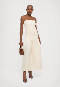 Trägerloses, cremefarbenes Kleid mit einem gerafften Oberteil und einem fließenden, gerafften Rock. Das Model hält eine braune Handtasche und trägt transparente Sandalen mit Absatz.