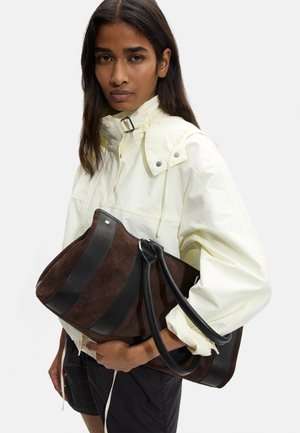 Kobieta w jasnożółtej kurtce i czarnych krótkich spodenkach trzyma dużą brązowo-czarną skórzaną torbę na jednolitym tle.