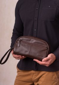Brown leather toiletry bag with a zipper, rounded shape, front pocket, and a short handle. Smooth texture and minimalistic design.
