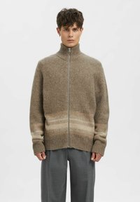 Vīrietis, kurš valkā brūnu vilnas džemperi ar rāvējslēdzēju, augstu apkakli un smalkām horizontālām svītrām, kombinācijā ar pelēkām biksēm, stāvot pret vienkāršu fona sienu.