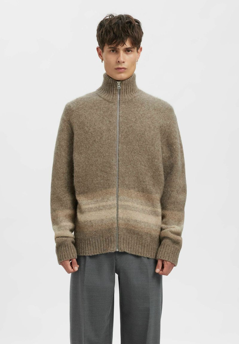 Vīrietis, kurš valkā brūnu vilnas džemperi ar rāvējslēdzēju, augstu apkakli un smalkām horizontālām svītrām, kombinācijā ar pelēkām biksēm, stāvot pret vienkāršu fona sienu.