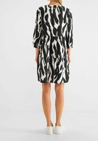 Žena v černo-bílých pruhovaných šatech po kolena s třičtvrtinovými rukávy a bílými teniskami, stojící čelem k tomu, co je za ní.