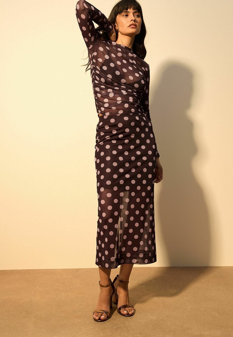 Robe sombre transparente à gros pois lilas, manches longues et silhouette ajustée. Assortie à des talons à brides de couleur similaire.