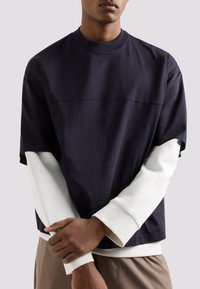 Top corto blu navy con scollo a giro, sovrapposto a una camicia a maniche lunghe bianca; presenta un dettaglio di cucitura piatta sul petto.