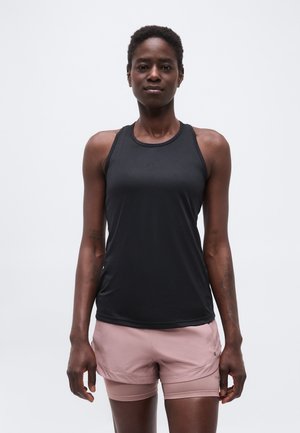 Femeie atletică purtând un top negru fără mâneci și pantaloni scurți roz deschis, stând în picioare pe un fundal simplu și deschis, cu fața spre înainte.