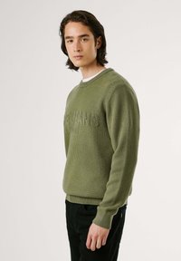 Žalias megztinis su apvalia iškirpte ir tekstūruotu raštu, su logotipu priekyje. Derinamas su juodomis kelnėmis.