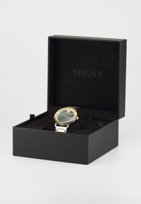 Reloj Versace con una esfera redonda verde y dorada, correa de acero inoxidable plateado y bisel detallado, presentado en una caja de exhibición negra.