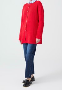 Red ribbed cardigan with buttons, worn over a blue and white striped collared shirt. Paired with dark blue straight-leg jeans and black loafers.