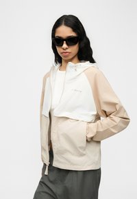 Νέα γυναίκα που φοράει γυαλιά ηλίου, μπεζ και λευκό μπουφάν Columbia, λευκό πουκάμισο και σκούρο γκρι παντελόνι, στέκεται με το ένα χέρι στην τσέπη.