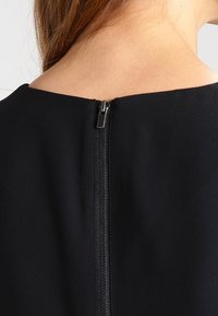 Vêtement noir avec une fermeture éclair au dos, présentant une texture lisse et mate ainsi qu'un design minimaliste. La fermeture éclair est de couleur argentée.