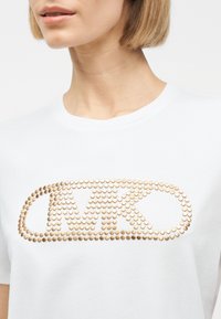 Λευκό πουκάμισο με χρυσούς κυκλικούς τρουκς που σχηματίζουν τα γράμματα "MK" μέσα σε ένα στρογγυλεμένο ορθογώνιο, φορεμένο από ένα άτομο με κοντά ξανθά μαλλιά.