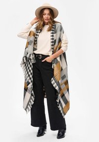 Szary, czarny i musztardowy szalik w kratę z frędzlami, noszony na jasnym swetrze i czarnych spodniach, w połączeniu z czarnymi botkami za kostkę.