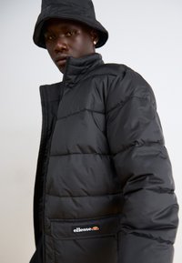 La giacca a vento nera presenta un design trapuntato, un colletto alto e una patch con logo. Completata da un cappello a secchiello nero coordinato. Texture liscia e lucida.