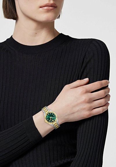 Femme portant un pull côtelé noir, les bras croisés, montrant une montre bicolore or et argent avec un cadran vert à son poignet gauche.