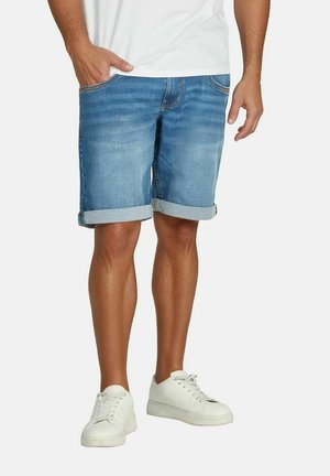 Mann trägt hochgekrempelte blaue Jeansshorts, weiße Sneakers und ein weißes T-Shirt, steht mit einer Hand in der Tasche.