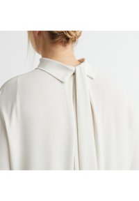 Weiße Bluse mit klassischem Kragen und einem drapierten Rückendetail. Glatte Textur mit einem weichen, fließenden Gewebe.