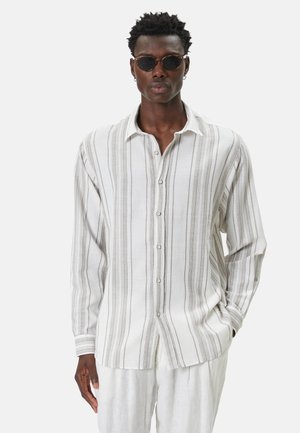 Mand iført runde solbriller og en hvid langærmet skjorte med lodrette grå striber, står med den ene hånd i lommen.