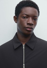 Black zip-up jacket with a pointed collar and visible metal zipper details, featuring a smooth, matte texture and minimalistic design.