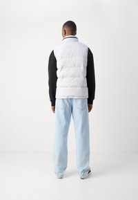 Gilet blanc matelassé avec un col haut sur une chemise à manches longues noire, porté avec un jean bleu clair à coupe ample. Coupe sur mesure, texture lisse.