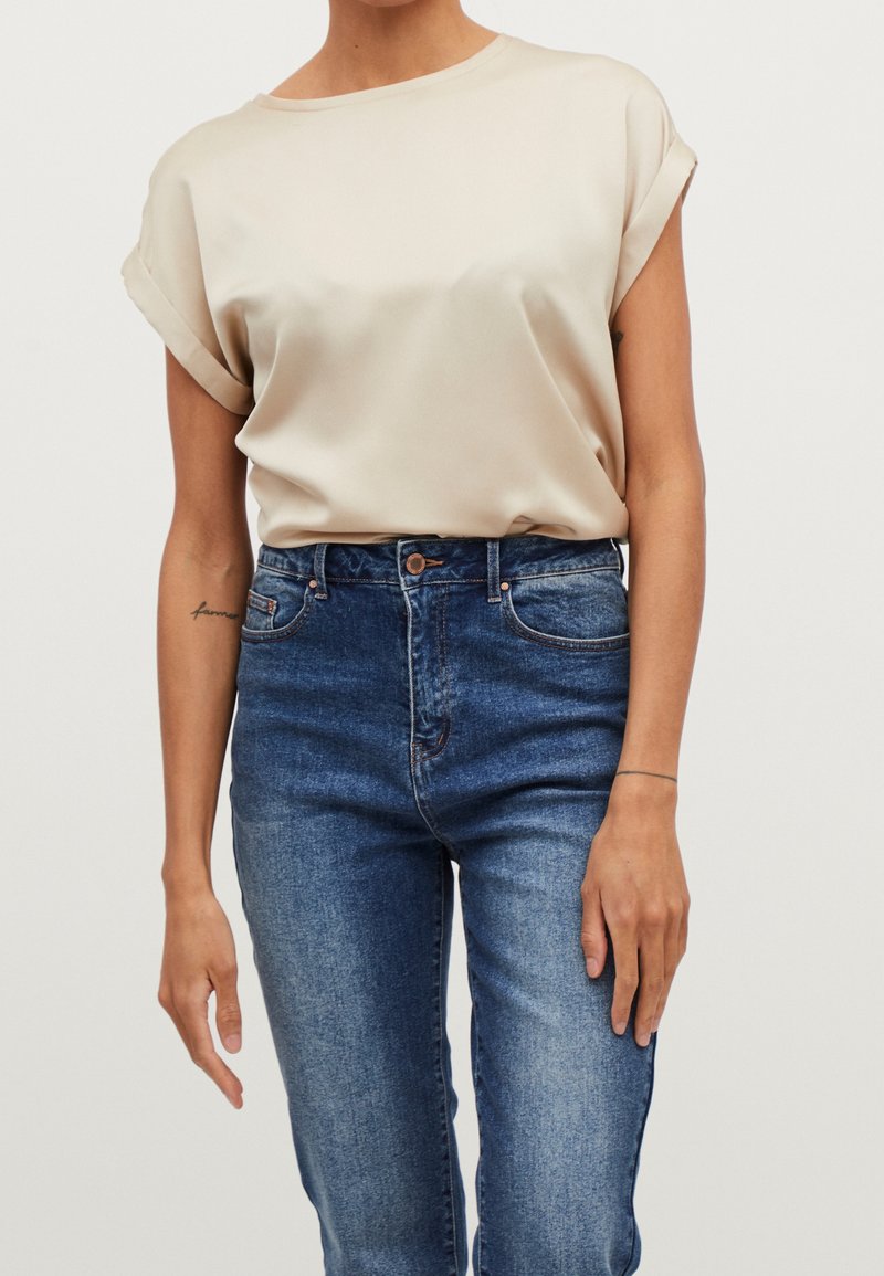Personne portant un chemisier beige à manches courtes avec des revers roulés, rentré dans un jean bleu taille haute sur un fond uni.