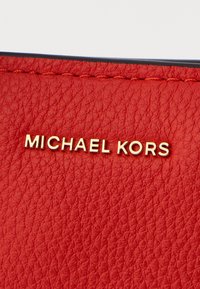 Pelle rossa testurizzata con branding in tonalità oro "MICHAEL KORS". La superficie presenta una finitura a granuli e cuciture a contrasto.