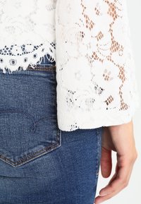 Haut en dentelle blanche avec un motif floral, présentant un ourlet effiloché, porté sur un jean en denim bleu foncé, avec une main reposant sur la hanche.
