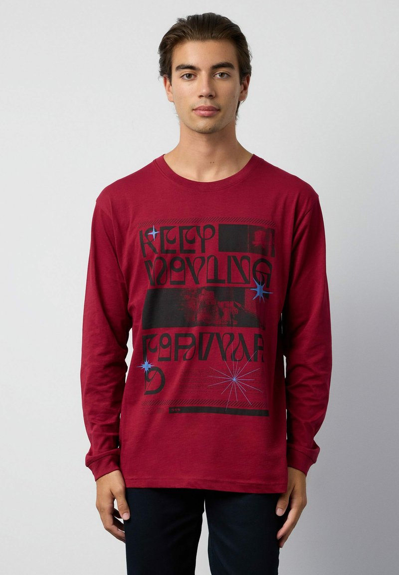 Joven wearing a red long-sleeve shirt with black and blue graphic text and star designs, standing against a plain light gray background.