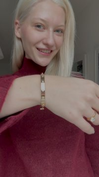 Kuldne käevõru, mille keskel on ristkülikukujuline charm, vahelduvate siledate ja tekstuuriga metallosadega. Kasutaja kannab burgundia värvi sviitrit.